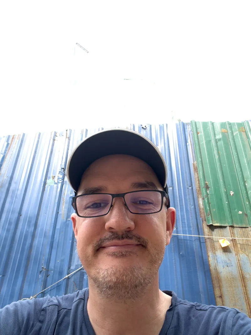 A man taking a selfie from a slightly low angle, looking directly at the camera with a subtle smile. He is wearing black-framed rectangular glasses, a black baseball cap, and a navy blue t-shirt. He has a goatee and mustache with light stubble. Behind him is a wall made of corrugated metal sheets, predominantly blue with a section of green and some rust patches visible. A thin wire or clothesline stretches horizontally across the background. The sky above the metal wall is bright and overcast, completely white.