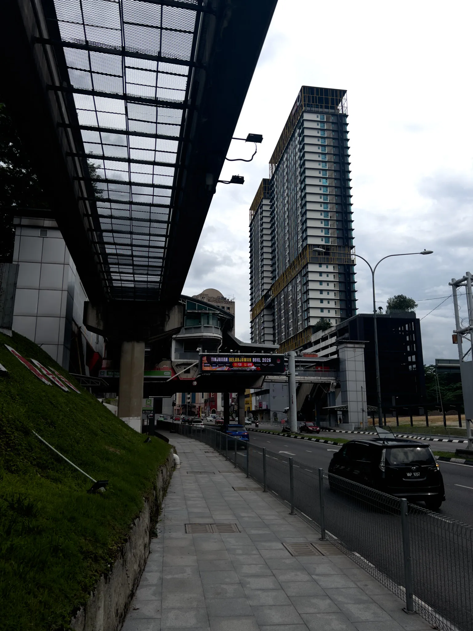 A covered pedestrian walkway runs alongside a multi-lane road in Kuala Lumpur, Malaysia. The walkway is paved with gray tiles and bordered by metal railings on the road side and a grassy, sloped embankment on the left. Above, a metal-and-mesh canopy provides shelter. In the background, a tall residential high-rise condominium tower with yellow and blue accents rises against an overcast sky. A monorail or elevated rail station with a distinctive domed roof structure is visible in the mid-ground, with elevated tracks and a pedestrian bridge crossing overhead. A digital banner on the bridge reads "Tinjauan Belanjawan DBKL 2026." Several vehicles, including a black car with a Malaysian license plate, travel along the road. Street lights, traffic infrastructure, and commercial buildings line the urban streetscape.