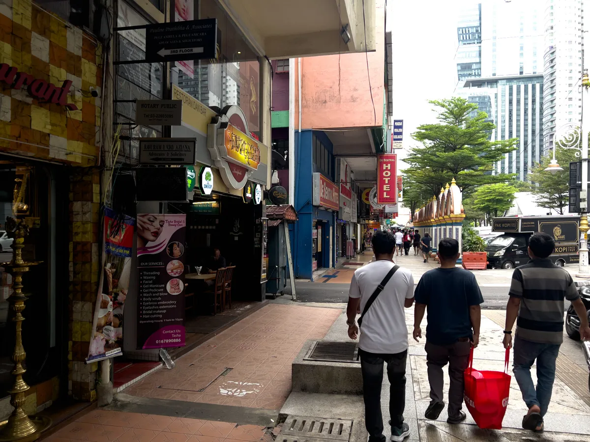 A busy street in Seputeh, Kuala Lumpur, 50614, Malaysia. In the foreground, three men walk along the grey paved sidewalk. Two wear T-shirts; the third carries a red bag. Numerous shops line the left side, including a restaurant and a beauty salon whose offers are advertised on a large poster. A hotel stands on the right, while modern office buildings reach toward the sky in the background. Trees and a decorative fence run along the street.