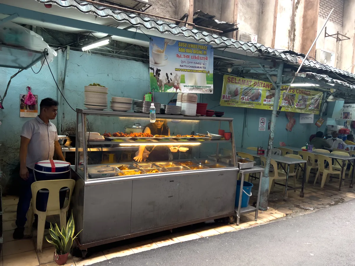 In Brickfields, Kuala Lumpur, stands a simple, inviting street restaurant. A counter displays various dishes, including meats and side dishes presented in several containers. A man in a shirt watches the guests while a woman at the stall prepares the food. Several signs above the stand show the menu in Malay. Plastic chairs and tables in the background offer seating. The surroundings are full of colour and life, reflecting the local food culture.