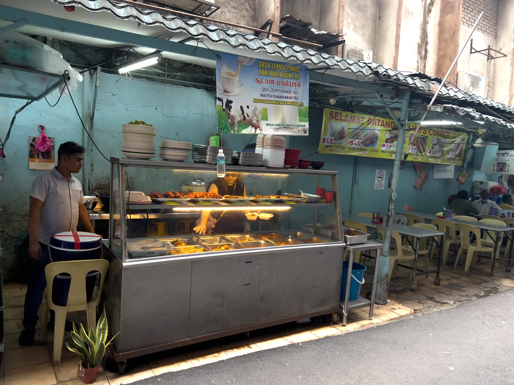 A Malaysian street food stall called "Sami's Corner" with a stainless steel buffet-style food display case filled with various cooked dishes including curries and fried items. A man in a white striped shirt stands to the left near stacked plates and bowls, while a woman behind the counter reaches into the display case. Signs in Malay, Tamil, and English advertise hot cow milk, Nattu Chakkarai Tea, and meal prices in Ringgit Malaysia. A yellow banner reads "Selamat Datang" (Welcome). The open-air eatery has a corrugated metal roof, light blue painted walls, plastic chairs and tables where diners are seated in the background, and a small potted snake plant near the front. A cooler box sits on one of the plastic chairs beside the stall.