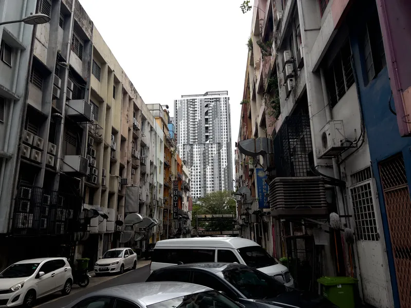 A narrow back alley in a Southeast Asian city, likely Kuala Lumpur, Malaysia, flanked by aging low-rise shophouse buildings on both sides. The buildings are weathered with visible stains, exposed air conditioning units, metal grilles on windows, and satellite dishes mounted on walls. On the left side, a small "Hotel" sign is visible among the colorful but worn facades in shades of cream, yellow, and orange. On the right side, a blue sign reading "Spektrum" is partially visible. The alley is congested with parked cars and a white van, leaving little room for passage. In the background, towering modern high-rise residential condominiums rise dramatically above the old buildings, creating a stark contrast between old and new urban development. The sky is overcast and grey, adding to the gritty atmosphere of the scene. Green trash bins and scattered vegetation on upper floors add small details to the cluttered urban landscape.