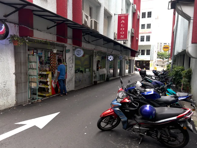 A narrow back alley in a Malaysian town lined with small shops and businesses. On the left side, a man in a blue t-shirt stands at the entrance of a convenience store with a green display rack of goods outside. Above the shop, a red vertical sign reads "Kedai Gunting Rambut Saloon" (a Malay barbershop sign). The buildings have red and white painted facades with metal awnings extending over the shopfronts. On the right side of the alley, a row of motorcycles and scooters are parked closely together, with a prominent red and black motorcycle in the foreground bearing a Malaysian license plate. A white directional arrow is painted on the asphalt road pointing forward. In the background, more buildings, signage including what appears to be a backpackers hostel and a tea cafe, and taller apartment blocks are visible. Potted plants and hanging greenery add touches of life to the urban setting.