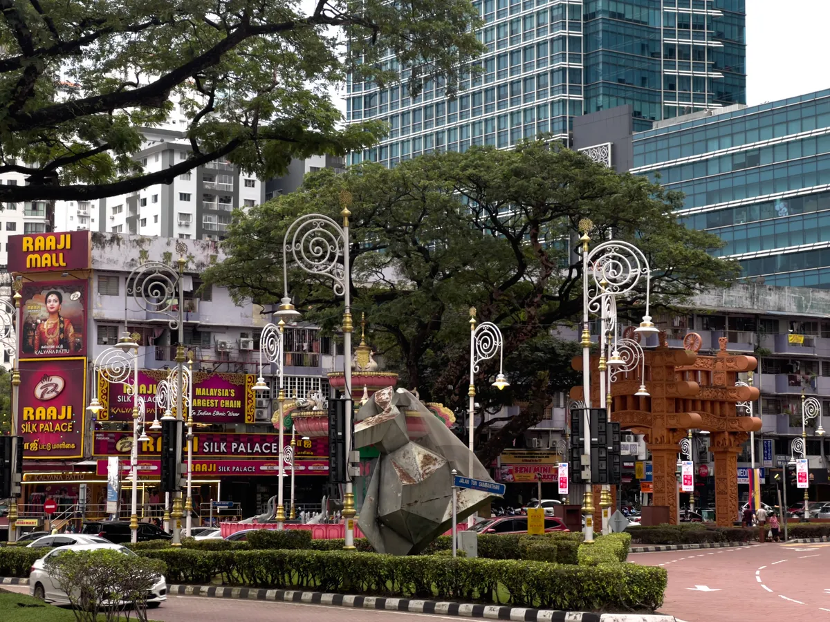 In Brickfields, Kuala Lumpur, a lively cityscape unfolds with colourful signs and advertisements for Raaji Mall and Raaji Silk Palace in the foreground. A modern, multi-storey building complex dominates the background. Colourful lanterns and a striking, ornate wooden structure blend harmoniously into the scene. Traffic signs and bicycles are part of the streetscape, while lush trees cast shade and add a touch of green to the urban space.