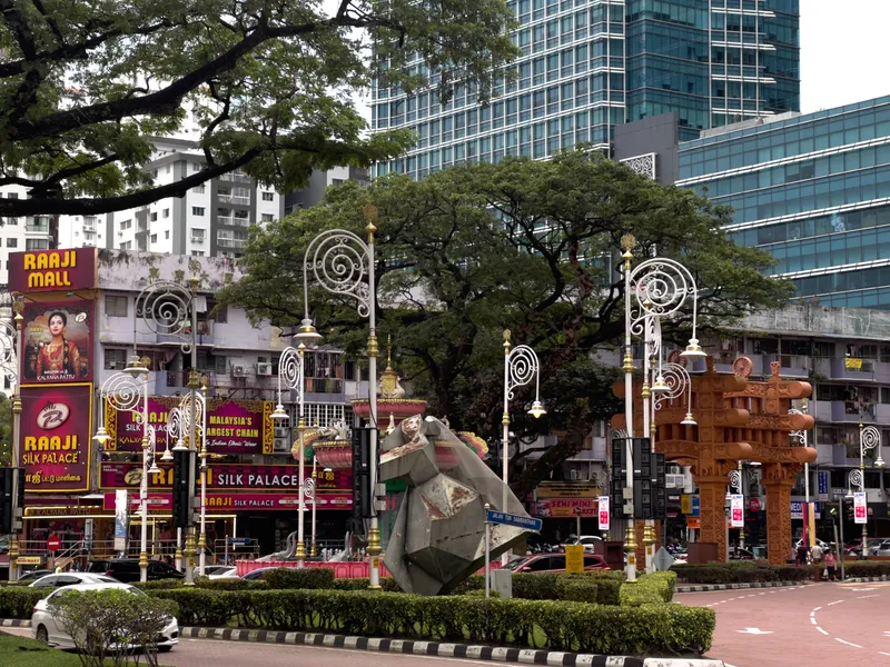 A bustling street scene in Little India, Kuala Lumpur, Malaysia, featuring a landscaped roundabout with a large geometric metallic sculpture at its center, surrounded by neatly trimmed hedges. Ornate white wrought-iron lampposts with gold accents and spiral designs line the area. On the left, colorful signage for "Raaji Mall" and "Raaji Silk Palace" advertises "Malaysia's Largest Chain in Indian Ethnic Wear" with vibrant pink and magenta billboards featuring a woman in traditional Indian attire. To the right stands a tall brown carved wooden or terracotta-style decorative pillar with traditional Indian architectural motifs. Large tropical rain trees with spreading canopies provide shade over the scene. In the background, modern glass-fronted high-rise buildings and older residential apartment blocks create a contrast between old and new urban architecture. Cars are parked and moving along the street, and a street sign reading "Jalan Tun Sambanthan" is partially visible.