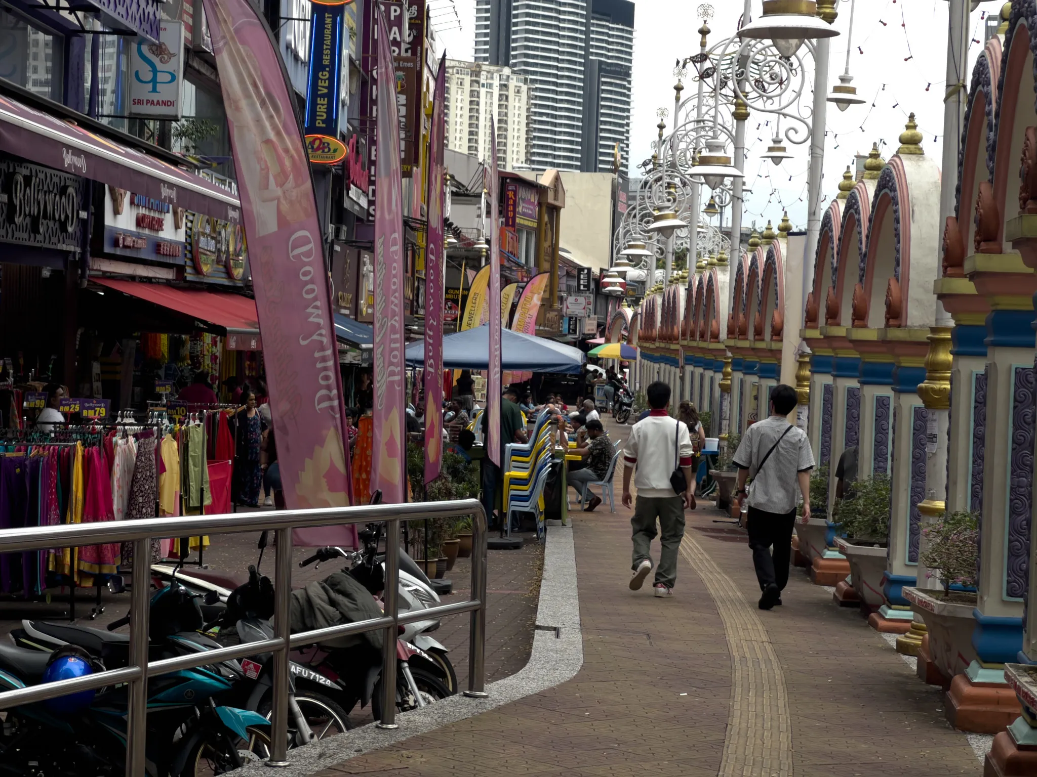 A bustling pedestrian walkway in Little India, Kuala Lumpur, Malaysia, flanked on the left by colorful shopfronts including "Sri Paandi" pure vegetarian restaurant and "Bollywood" themed stores with pink feather banners. Racks of vibrant saris and clothing in yellow, pink, red, and purple line the sidewalk. On the right side, an ornate Hindu temple wall features decorative arches in cream, blue, gold, and terracotta colors, with elaborate ornamental streetlamps rising above. Several pedestrians walk along the brick-paved path, while motorcycles and scooters are parked behind metal railings in the foreground. Blue and yellow canopy tents shelter outdoor dining areas in the middle distance. High-rise buildings and skyscrapers are visible in the background against an overcast sky, creating a striking contrast between the traditional cultural district and the modern city skyline.