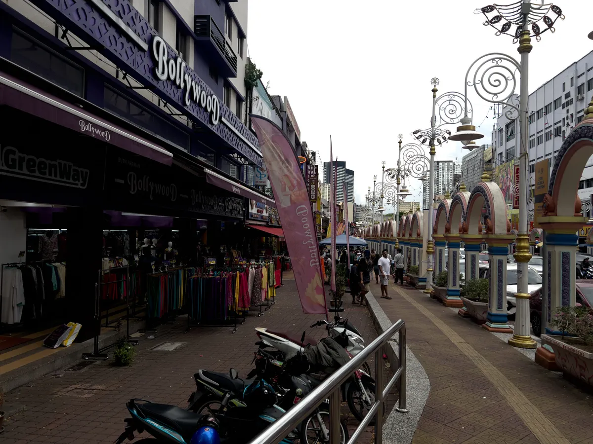 A lively shopping street lined with colourful stalls and shops runs through Brickfields, Kuala Lumpur, 50470, Malaysia. On the left stands a shop called 'Bollywood', stocked with vibrant clothing on display racks. The pavements are framed by a curved railing and several motorbikes are parked along them. On the right, a row of atmospheric street lamps and decorative arches adds to the neighbourhood's cultural feel. The scene is busy with passers-by bringing it to life.