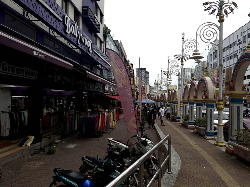 A bustling street in Little India, Kuala Lumpur, Malaysia, featuring colorful shopfronts including a prominent purple "Bollywood" clothing store with mannequins and racks of vibrant garments displayed on the sidewalk. The pedestrian walkway is lined with ornate decorative arches in pastel colors of cream, blue, and gold, alongside elaborate wrought-iron street lamps with spiral designs. Motorcycles are parked behind a metal railing in the foreground. Several pedestrians walk along the wide sidewalk, with modern high-rise buildings visible in the background against an overcast sky. A pink feather banner flutters near the shop entrance, and additional retail stores stretch down the left side of the street. Cars are visible on the road to the right beyond the decorative archways.