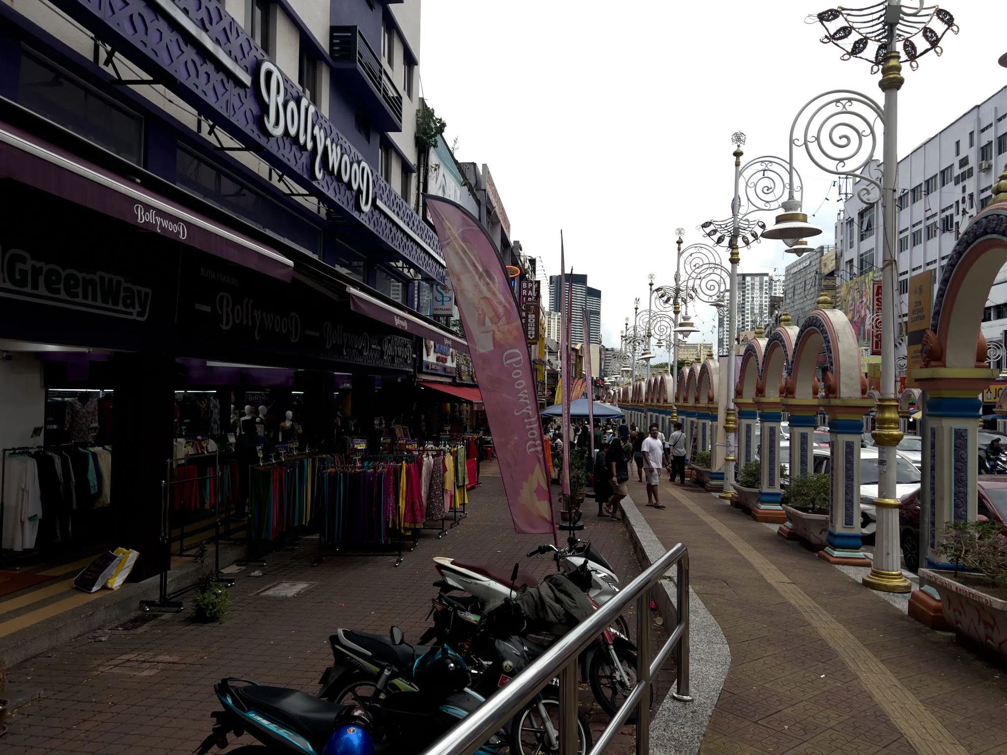 A bustling street in Little India, Kuala Lumpur, Malaysia, featuring colorful shopfronts including a prominent purple "Bollywood" clothing store with mannequins and racks of vibrant garments displayed on the sidewalk. The pedestrian walkway is lined with ornate decorative arches in pastel colors of cream, blue, and gold, alongside elaborate wrought-iron street lamps with spiral designs. Motorcycles are parked behind a metal railing in the foreground. Several pedestrians walk along the wide sidewalk, with modern high-rise buildings visible in the background against an overcast sky. A pink feather banner flutters near the shop entrance, and additional retail stores stretch down the left side of the street. Cars are visible on the road to the right beyond the decorative archways.