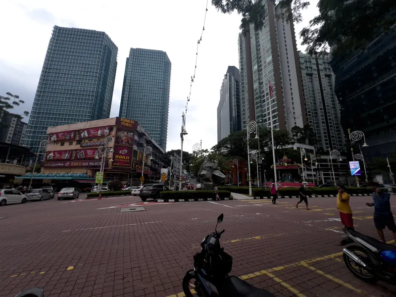 A bustling urban intersection in Kuala Lumpur's Little India (Brickfields area), featuring the colorful Raaji Mall and Raaji Silk Palace buildings with vibrant signage advertising Kalyana Pattu sarees on the left side. Tall modern glass skyscrapers and high-rise residential towers dominate the skyline in the background, contrasting with the older low-rise commercial buildings. A central median with manicured hedges and a modern abstract sculpture separates the lanes, while decorative spiral-topped street lamps line the road. Pedestrians cross the brick-paved intersection, motorcycles are parked in the foreground, and vehicles navigate the streets. A Malaysian flag is visible on one of the lampposts. The overcast sky casts a diffused light over the scene, highlighting the mix of traditional Indian commercial culture and modern metropolitan development.