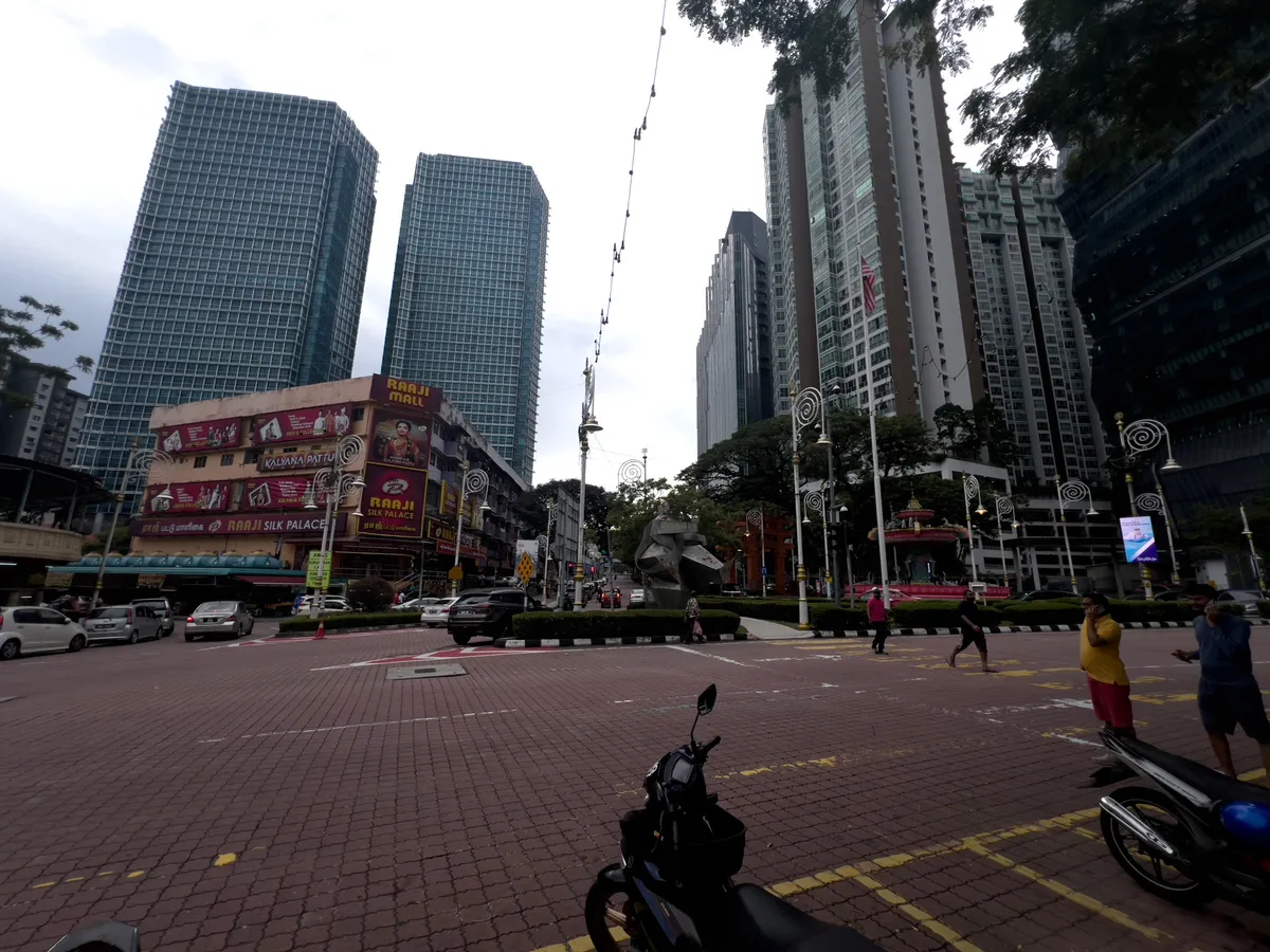 A bustling city scene in Brickfields, Kuala Lumpur. Several vehicles fill the paved road in the foreground while pedestrians cross the street. On the left rises the multi-storey 'Raji Mall' building, covered in colourful advertising posters. Modern high-rises tower into the sky in the background, their glass facades reflecting the surrounding trees and sky. Street lamps and decorations underline the urban character of the neighbourhood, while a lightly clouded sky hangs above the scene.