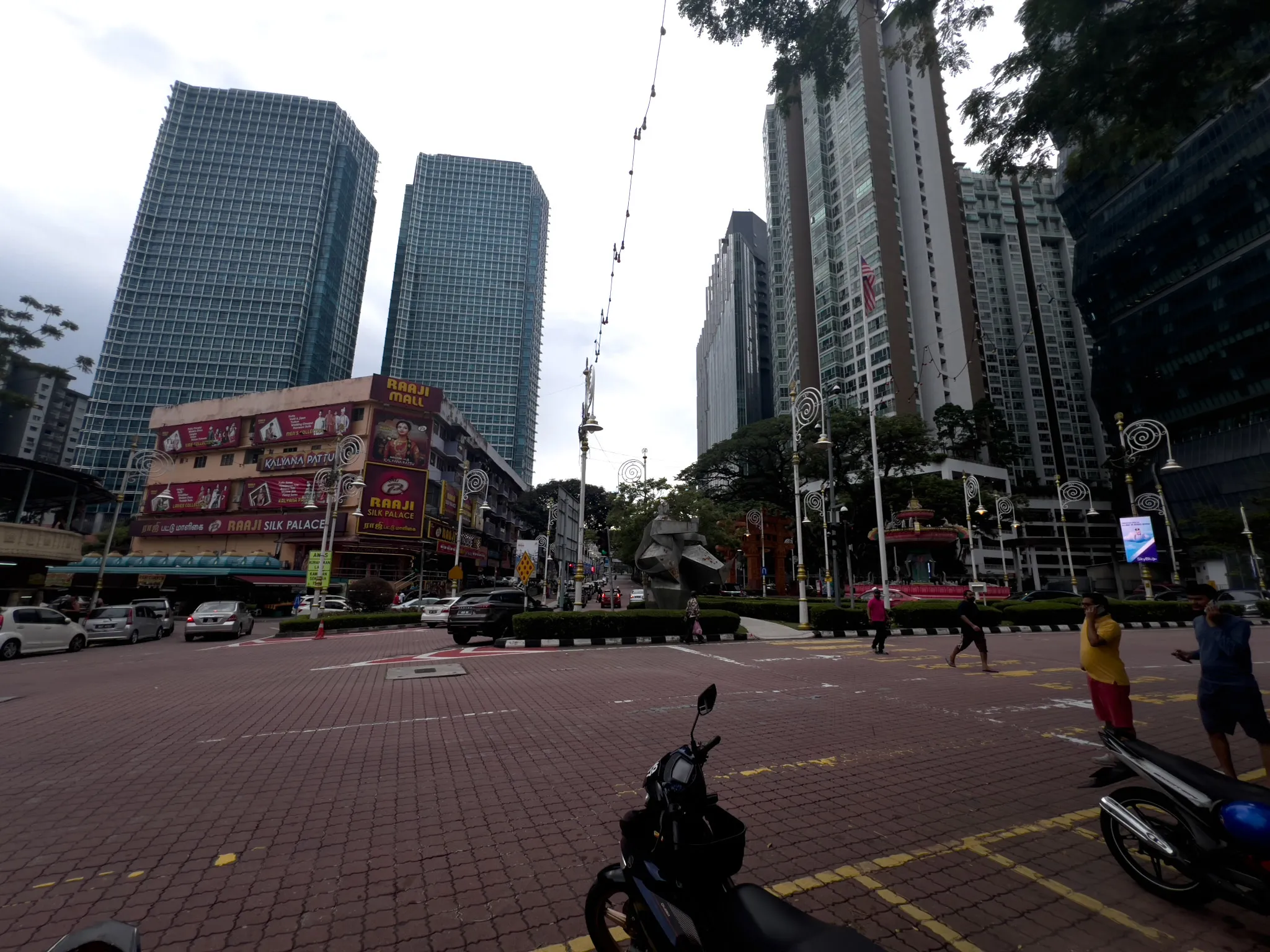 A bustling urban intersection in Kuala Lumpur's Little India (Brickfields area), featuring the colorful Raaji Mall and Raaji Silk Palace buildings with vibrant signage advertising Kalyana Pattu sarees on the left side. Tall modern glass skyscrapers and high-rise residential towers dominate the skyline in the background, contrasting with the older low-rise commercial buildings. A central median with manicured hedges and a modern abstract sculpture separates the lanes, while decorative spiral-topped street lamps line the road. Pedestrians cross the brick-paved intersection, motorcycles are parked in the foreground, and vehicles navigate the streets. A Malaysian flag is visible on one of the lampposts. The overcast sky casts a diffused light over the scene, highlighting the mix of traditional Indian commercial culture and modern metropolitan development.