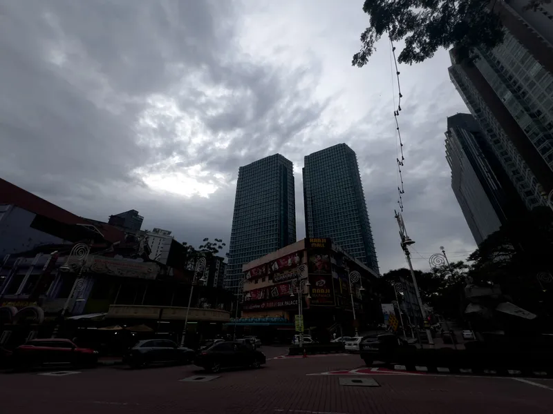 A dramatic overcast sky looms over a busy urban intersection in Kuala Lumpur's Little India district. Two tall glass skyscrapers rise prominently in the center background against heavy grey clouds with faint sunlight breaking through. At street level, colorful shopfronts line the road, including "Raaji Mall" and "Raaji Silk Palace" with vibrant signage and Bollywood-style advertisements. Ornamental street lamps with spiral decorative elements stand along the brick-paved sidewalks. Several cars are parked and moving along the road. Trees frame the upper right corner, and a string of hanging lights stretches across the scene. The low-light conditions give the entire scene a moody, dusk-like atmosphere with deep shadows at street level contrasting against the luminous cloud cover above.
