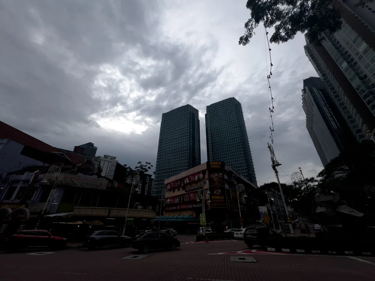 Two ultra-modern skyscrapers clad in dark glass rise in the background of Brickfields, Kuala Lumpur. Grey clouds cover the sky, creating a sombre atmosphere. In the foreground, several vehicles move along the street amid busy city life. Colourful buildings with signs and lights advertising shops line the left side. A tall sculptural element also stands in the scene, reinforcing the urban character of the surroundings.
