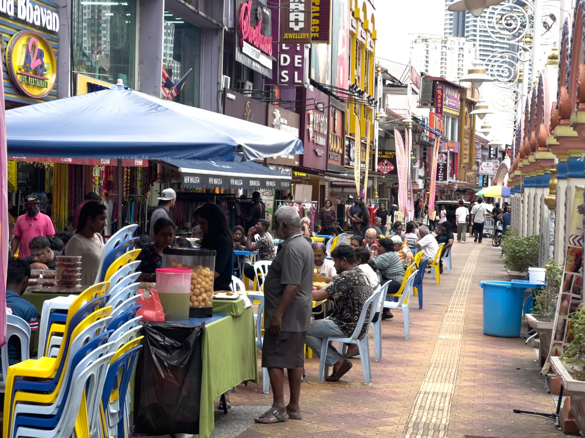 Rows of blue and yellow chairs line a paved walkway in Brickfields, Kuala Lumpur, 50470, Malaysia. At a stall, vendors under a blue tent prepare various dishes while people sit at plastic tables eating and drinking. Colourful shops with signage are visible in the background. People stroll along the street and the atmosphere is lively.