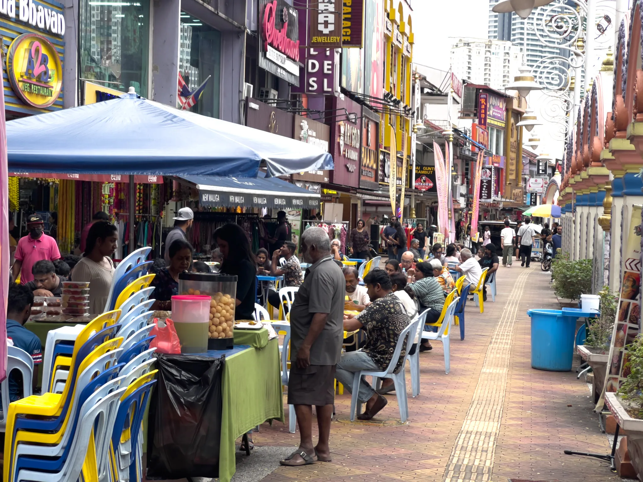 A bustling pedestrian street in Little India, Kuala Lumpur, Malaysia, lined with colorful shopfronts displaying signs for restaurants like "Bhavan" and "A2B Veg Restaurant," jewellery stores, and textile shops. Blue and yellow plastic chairs are arranged along outdoor dining areas under a blue canopy tent, where locals sit eating street food. A street vendor stands near a food cart displaying pani puri (golgappa) in a glass container. The brick-paved walkway stretches into the distance, flanked on the right by ornate Hindu temple architecture with blue and gold decorative elements. Tall modern buildings rise in the background, contrasting with the traditional shophouses. Pedestrians walk along the lane, and vibrant banners and signage in multiple languages add to the lively, multicultural atmosphere. Stacked chairs in blue and yellow sit ready for more customers along the busy food street.