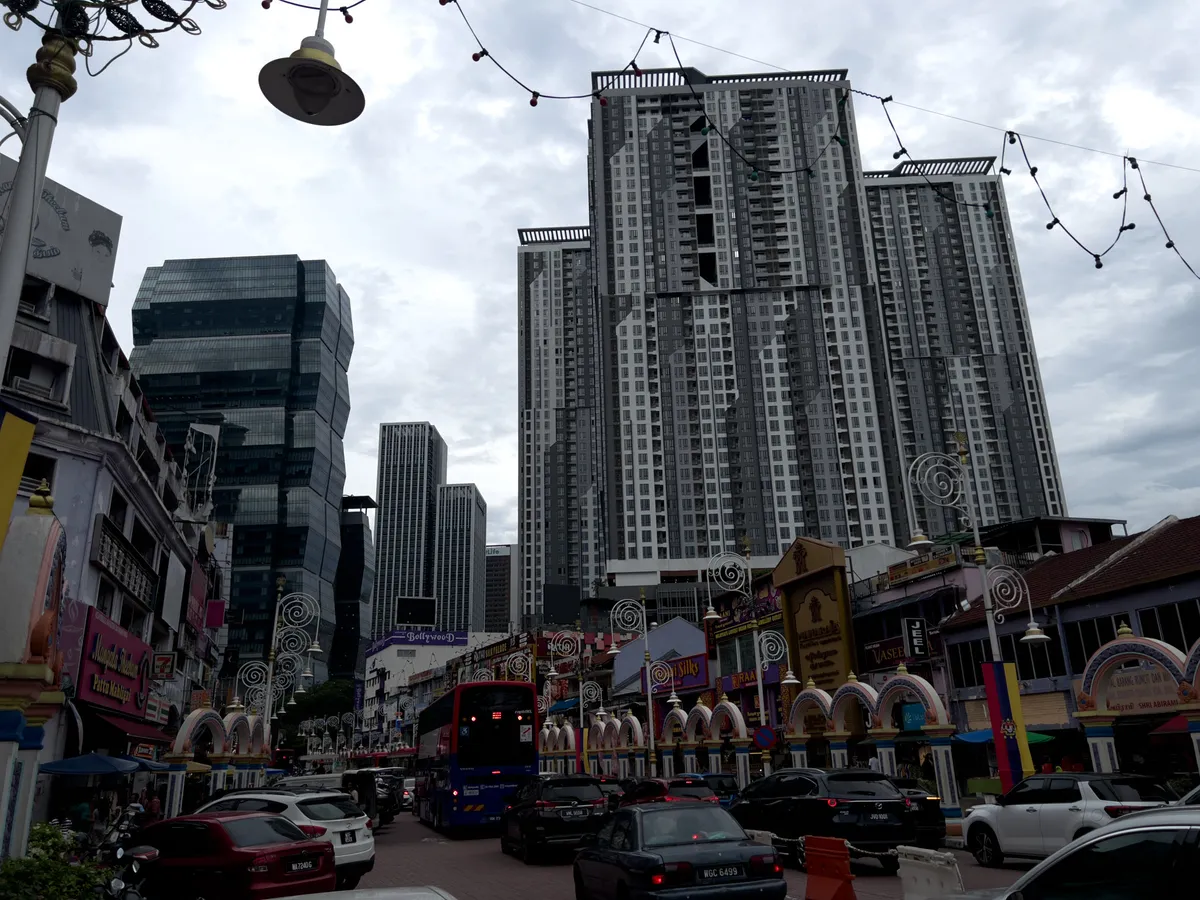 A busy street runs through Brickfields, Kuala Lumpur, 50470, Malaysia, with colourful shops and facades in the foreground. Multi-storey, historic-looking buildings stand on the left, while on the right modern high-rises, including a striking twisted tower, rise into the sky. A red city bus drives along the street and several cars are parked along the approach. Decorative lights hang above the scene and a grey, cloudy sky enhances the urban atmosphere.