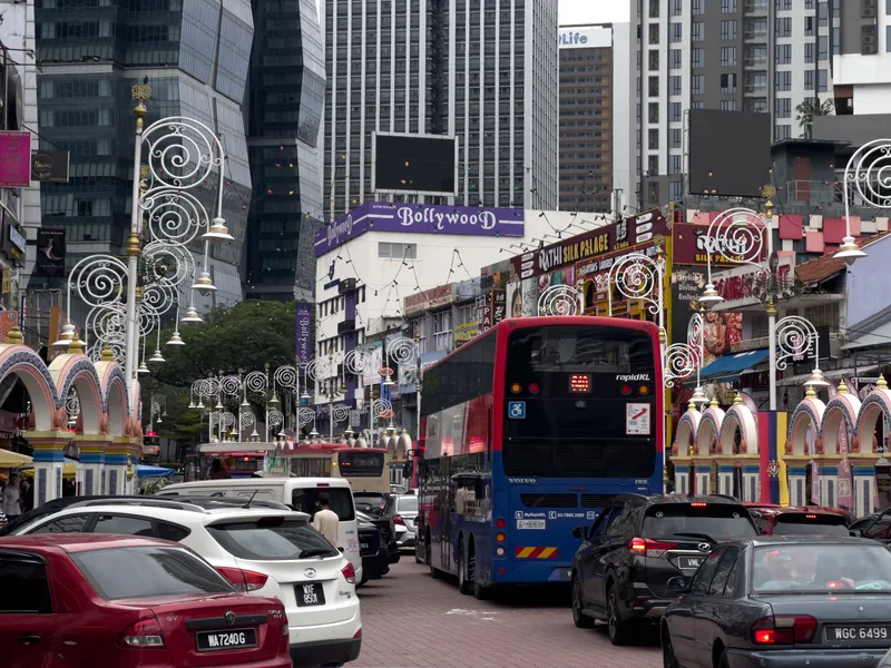 A busy street scene in Little India, Kuala Lumpur, Malaysia, featuring a red and blue RapidKL Volvo double-decker bus navigating through heavy traffic. The street is congested with numerous cars, including vehicles with Malaysian license plates. On both sides of the road, colorful Indian-style archways with pastel pink, yellow, and blue decorations frame the scene. Ornate white wrought-iron street lamps with spiral designs and golden accents line the median. Prominent shop signs include "Bollywood" in purple and "Rathi Silk Palace" in red, indicating the area's South Asian cultural character. In the background, modern high-rise buildings and skyscrapers tower over the low-rise commercial shophouses, creating a contrast between traditional ethnic neighborhood character and contemporary urban development. The overcast sky adds a muted tone to the vibrant streetscape.