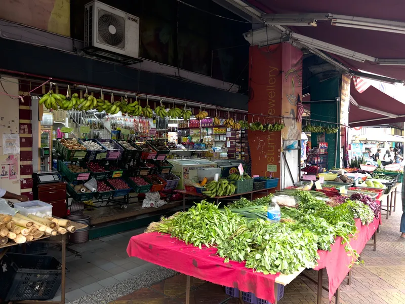 An outdoor street market stall in what appears to be a Malaysian neighborhood, likely Little India, displaying a vibrant array of fresh produce. In the foreground, a large table covered with a pink cloth is piled high with leafy green vegetables including kangkung (water spinach), pandan leaves, and other Asian greens. Behind the table, a storefront features hanging bunches of green bananas suspended from hooks along the top of the shop entrance. Below the bananas, plastic crates are filled with onions, shallots, potatoes, garlic, and other root vegetables. Sugarcane stalks are bundled on the left side. The shop interior is stocked with bottled goods and packaged products. A red-brown pillar with gold text reading "Costume Jewellery" separates this stall from the adjacent shops. A Malaysian flag is visible on the pillar. The covered walkway extends to the right where more vegetable stalls and pedestrians can be seen in the background. A York air conditioning unit is mounted above the storefront.