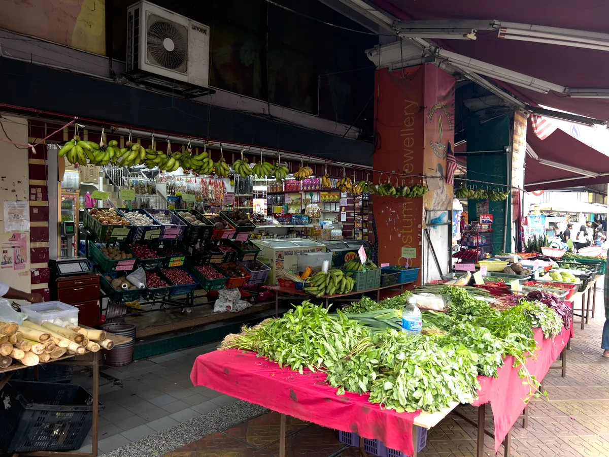 A lively market spreads through Brickfields, Kuala Lumpur, offering all kinds of fresh produce. In the foreground, tables display a variety of fresh herbs and vegetables on red cloths. On one table are long stems and fresh greens. Shelves of fruit and other goods stand in the background while bunches of bananas hang above the market. The scene is busy and captures the market's lively bustle.