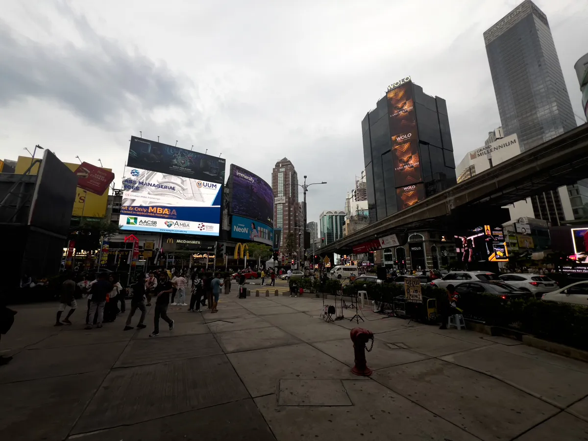 A bustling cityscape unfolds in Kampung Cendana, Kuala Lumpur. In the foreground lies a large square with numerous people moving about and lingering. High-rises with glowing billboards define the skyline, while the sky is covered in grey clouds. A large screen displays advertising, and a McDonald's outlet is visible nearby. Cars travel along the street, and the modern architecture of the buildings gives the scene a distinctly urban character.