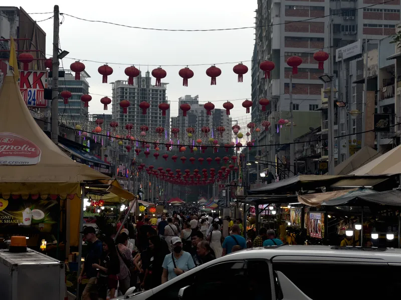 A bustling street market in Kuala Lumpur's Chinatown (Petaling Street) at dusk, with rows of red Chinese lanterns strung across the street between buildings. The narrow street is packed with crowds of people walking between food stalls and vendor canopies on both sides. On the left, a yellow tent displays a Magnolia ice cream sign and a "KK 24 Jam" convenience store sign is visible. On the right, The Quay Hotel sign can be seen on one of the multi-story buildings. High-rise apartment buildings and construction sites are visible in the background against an overcast sky. Street food vendors with illuminated stalls line both sides, while colorful decorative lanterns in red, yellow, pink, and green hang at varying heights. A white vehicle is parked in the foreground on the right side, and the scene captures the vibrant energy of a Southeast Asian night market coming alive as evening approaches.
