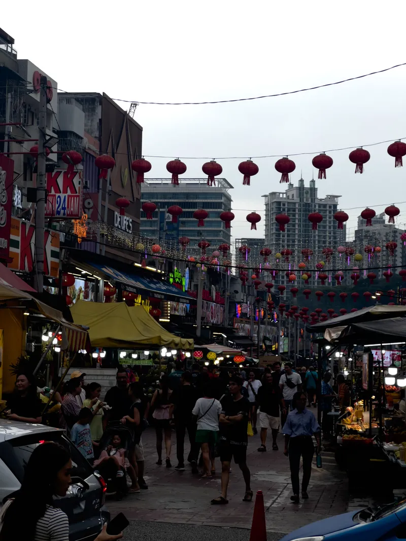 A bustling street market in Kuala Lumpur's Chinatown (Petaling Street) during dusk, with rows of red Chinese lanterns strung across the street overhead. Crowds of pedestrians walk along the narrow pedestrian area flanked by vendor stalls with yellow and striped canopies on both sides. Shop signs including "KK 24 JAM" and "DE LIFE'S" are visible on the left side of multi-story shophouses. In the background, modern high-rise buildings and construction can be seen against an overcast sky. Colorful neon lights and string lights illuminate the market area, while food stalls and merchandise vendors line both sides of the street. A traffic cone marks the edge of the pedestrian zone in the foreground, where a parked car and motorcycle are also visible. People of various ages, including families with young children, browse and walk through the lively night market scene.