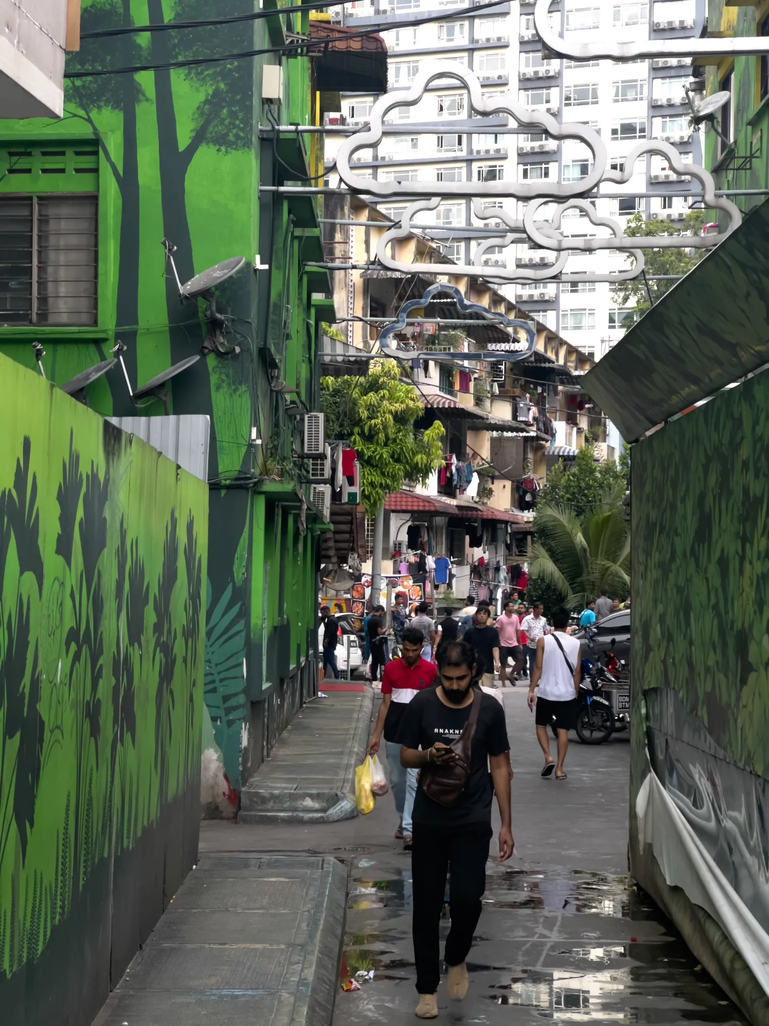 A narrow alleyway in a Southeast Asian urban neighborhood, flanked by walls painted with vibrant green murals depicting trees, tropical plants, and foliage. A man in a black t-shirt with a crossbody bag walks toward the viewer while checking his phone and carrying a plastic bag. Behind him, another person in a red and white shirt follows, with more pedestrians visible further down the busy lane. Overhead, decorative white cloud-shaped metal installations span across the alley. The background reveals densely packed residential apartment buildings with laundry hanging from balconies, satellite dishes, air conditioning units, and parked motorcycles along the street. Puddles of water dot the concrete walkway, suggesting recent rain. The scene captures the lively, layered atmosphere of a working-class urban community with artistic street beautification efforts.