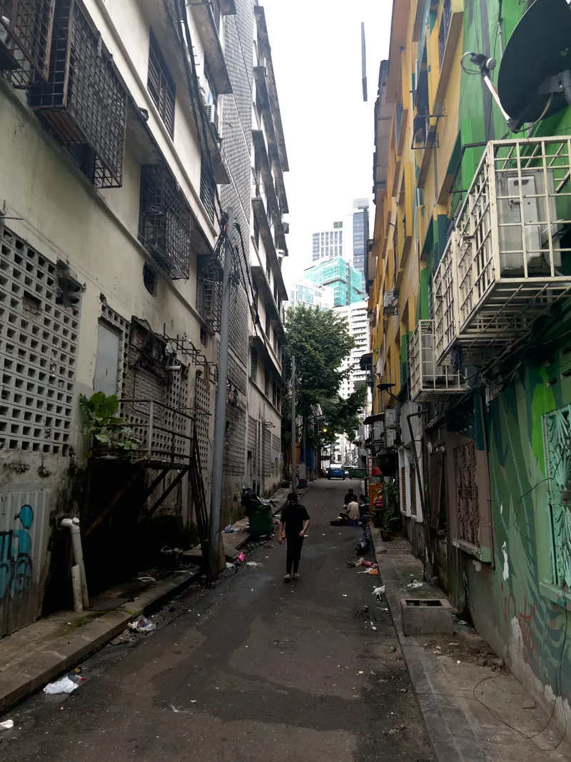 A narrow back alley in a Southeast Asian city, flanked by aging multi-story buildings on both sides. On the left, weathered concrete buildings with barred windows, decorative breeze block walls, and visible water stains show signs of urban decay. On the right, a building painted in vibrant green and yellow with colorful murals or graffiti contrasts with the drab surroundings, also featuring barred windows and a satellite dish. A person wearing a black shirt and cap walks away from the camera along the paved alleyway, which is littered with scattered trash and debris. Open drainage channels line both sides of the narrow lane. Further down, another person sits near the right-side buildings. In the background, a large green tree rises above the low-rise structures, and modern high-rise buildings and skyscrapers are visible against an overcast sky, highlighting the stark contrast between old neighborhood back lanes and the developing urban skyline. Utility poles, electrical wires, air conditioning units, and potted plants add to the cluttered, lived-in atmosphere of the scene.