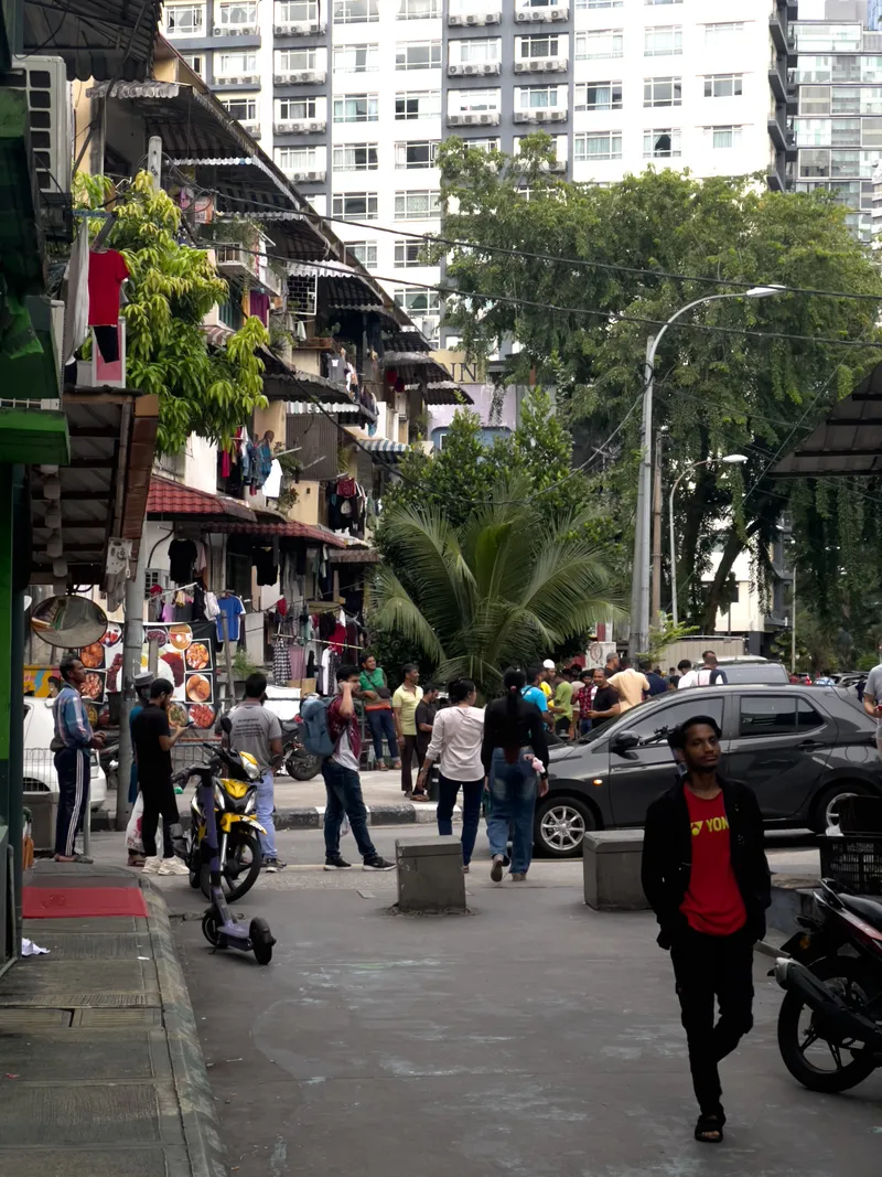A busy urban street scene in a Southeast Asian city, likely Kuala Lumpur, Malaysia. In the foreground, a man wearing a red Yonex t-shirt under a black jacket walks toward the camera. Behind him, numerous pedestrians mill about on the street and sidewalks. To the left, old shophouses with laundry hanging from balconies and food menu photographs displayed outside a restaurant line the street. A yellow motorcycle and an electric scooter are parked near the curb. Palm trees and other tropical greenery are visible in the middle ground. In the background, a modern high-rise apartment building with rows of air conditioning units contrasts sharply with the older low-rise shophouses in the foreground. Street lamps, power lines, and parked cars including a dark SUV add to the dense, lively atmosphere of this crowded neighborhood street.