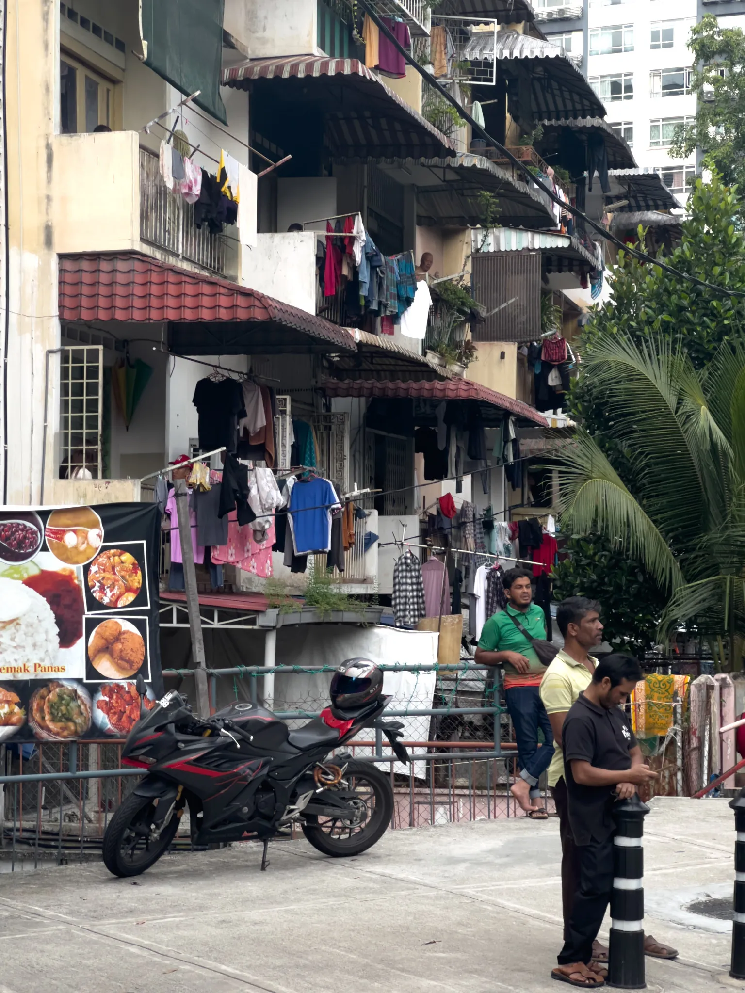 A bustling street scene in a Southeast Asian city, likely Malaysia, featuring a densely packed residential apartment building with multiple floors. Laundry hangs from nearly every balcony and railing, with colorful clothing draped on poles and lines across the facade. The building has a weathered, aged appearance with red-tiled awnings, corrugated metal overhangs, and cream-colored walls. At street level, a black sport motorcycle with a helmet resting on the seat is parked next to a metal railing. A food advertisement banner on the left displays images of local dishes including rice, curry, and a sign reading "Nasi Lemak Panas." Three men stand near the railing — one in a green polo shirt, one in a yellow shirt, and one in a dark shirt looking at his phone. Palm trees and tropical vegetation grow beside the building, and a modern high-rise is partially visible in the background, contrasting with the older residential block.