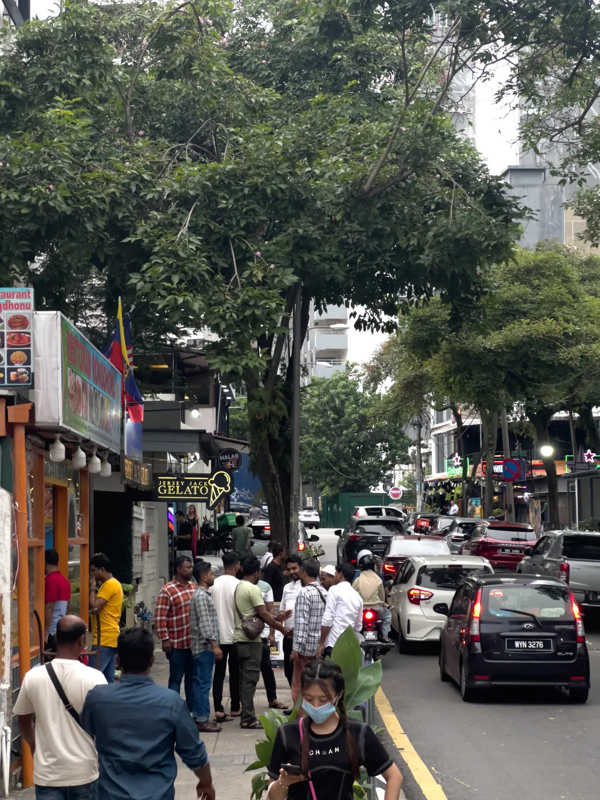 In Kampung Cendana, Kuala Lumpur, the scene shows a busy street with a mix of pedestrians and vehicles. Several groups of people gather on the pavement, chatting. Some wear casual clothes; one man is in a yellow T-shirt and another in a checked shirt. Cars and motorbikes move along the road in the background, while a 'Gelato' ice-cream parlour and another shop with colourful lights catch the eye. Green trees line the area, giving a shady feel, and it is a cloudy day.