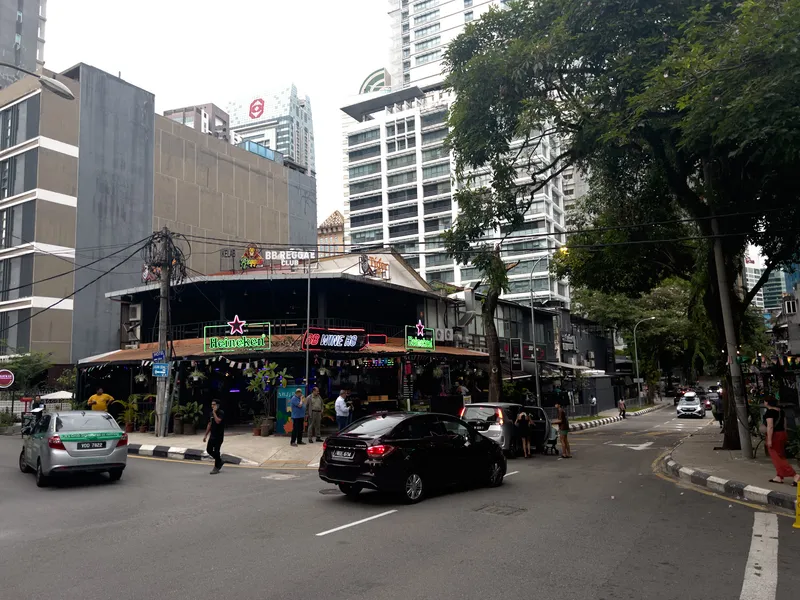 A busy urban street corner in Kuala Lumpur, Malaysia, featuring the BB Reggae Club and BB Wine HQ, open-air bars and nightlife venues adorned with neon signs including green Heineken logos. The low-rise entertainment establishments sit at a corner intersection, surrounded by tall modern high-rise buildings and skyscrapers in the background. Several cars are on the road, including a black sedan and a silver taxi with a green stripe. Pedestrians walk along the sidewalks and near the venue entrances. Lush tropical trees with dense green foliage line the right side of the street. Power lines and utility poles are visible, and the overcast sky suggests either dusk or an overcast day. The venue features tropical plants at its entrance, string lights, and colorful neon signage creating a vibrant nightlife atmosphere in the heart of the city.