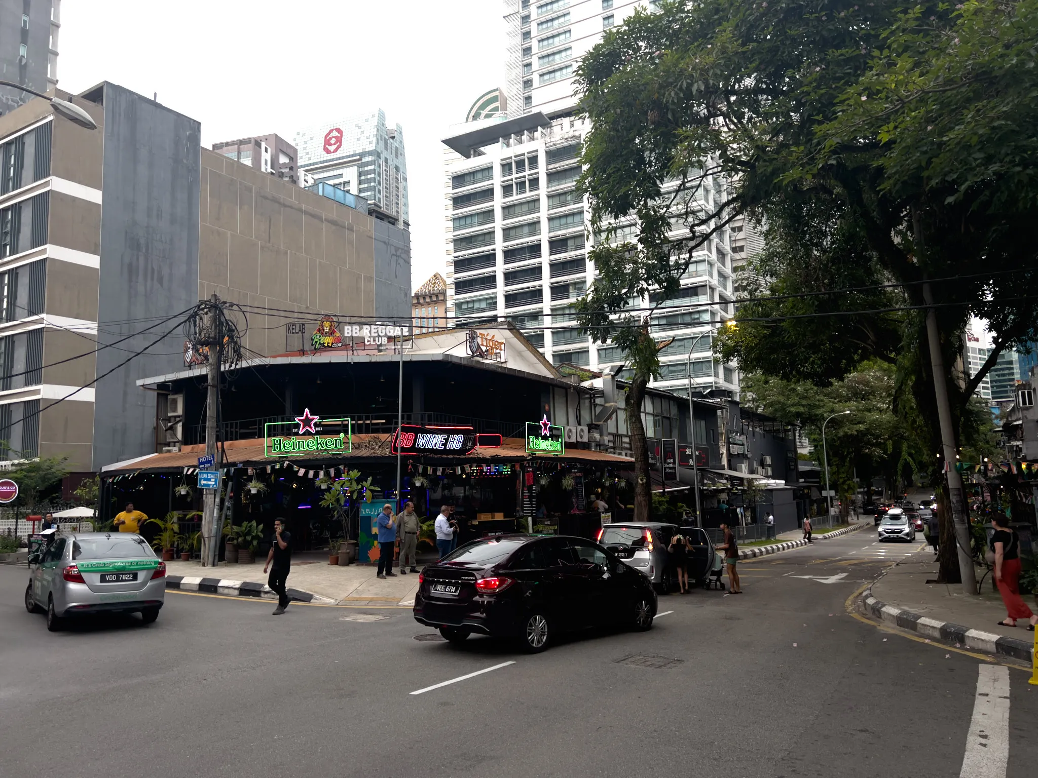 A busy urban street corner in Kuala Lumpur, Malaysia, featuring the BB Reggae Club and BB Wine HQ, open-air bars and nightlife venues adorned with neon signs including green Heineken logos. The low-rise entertainment establishments sit at a corner intersection, surrounded by tall modern high-rise buildings and skyscrapers in the background. Several cars are on the road, including a black sedan and a silver taxi with a green stripe. Pedestrians walk along the sidewalks and near the venue entrances. Lush tropical trees with dense green foliage line the right side of the street. Power lines and utility poles are visible, and the overcast sky suggests either dusk or an overcast day. The venue features tropical plants at its entrance, string lights, and colorful neon signage creating a vibrant nightlife atmosphere in the heart of the city.