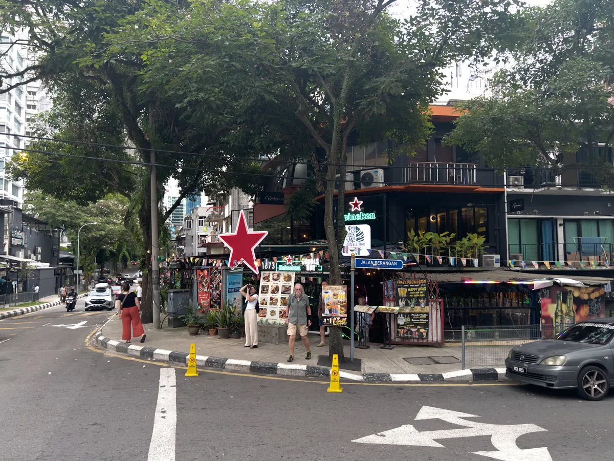 A busy street corner in Kampung Cendana, Kuala Lumpur, 50300, Malaysia. A large red star-shaped sign on the right marks a bar or restaurant. In the foreground, people walk along the street while a few cars and a motorbike pass by. Many stalls with colourful posters and advertising signs line the left side. The street is lined with trees that cast shade, and modern buildings are visible in the background.