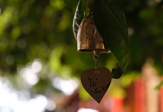 Eine kleine goldene Tempelglocke aus Bronze hängt an einem Ast zwischen grünen Blättern. An der Glocke baumelt ein herzförmiges Metallplättchen mit handgeschriebenen thailändischen Schriftzeichen, die einen Namen und ein Datum zu enthalten scheinen. Die Glocke ist kunstvoll verziert mit eingraviertem Relief, das eine Buddha-Figur darstellt. Im unscharfen Hintergrund sind üppiges grünes Laub und ein rötliches Gebäude erkennbar, das auf eine buddhistische Tempelanlage hindeutet. Weiches, durch das Blätterdach gefiltertes Sonnenlicht erzeugt ein sanftes Bokeh im Hintergrund.