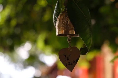 A small golden bronze temple bell hangs from a branch among green leaves. A heart-shaped metal plate dangles from the bell, bearing handwritten Thai characters that appear to contain a name and date. The bell is ornately decorated with engraved relief depicting a Buddha figure. In the blurred background, lush green foliage and a reddish building can be seen, suggesting a Buddhist temple complex. Soft sunlight filtered through the canopy creates a gentle bokeh in the background.