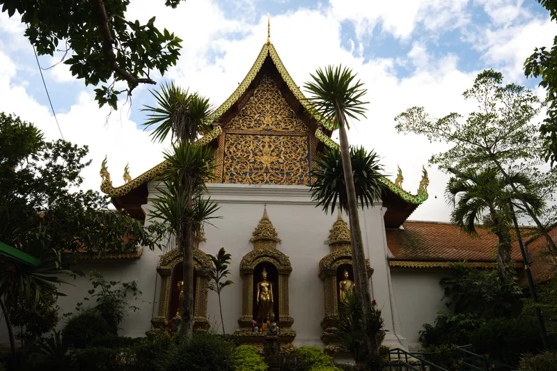A richly decorated Thai temple façade surrounded by lush tropical greenery. The central gable is adorned with elaborate gold and blue patterns, rising to a pointed peak with ornate serpent-like details along the roof edges. Below the gable, three golden Buddha statues stand in recessed niches framed by intricate gold carvings. Tall palm-like trees and dense foliage border the structure, while the sky above is partly cloudy and bright, creating contrast with the vivid architectural details.