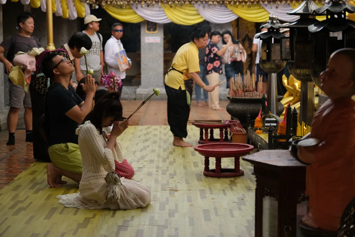 Visitors at a Buddhist temple in Thailand during worship. In the foreground, a young woman in a white dress and a man in a black T-shirt kneel on the tiled floor, both with hands folded in the prayer gesture (Wai). The woman holds incense sticks and a lotus blossom between her hands. On the right, golden monk statues, candles, flower offerings, and a large incense vessel with numerous burning incense sticks can be seen. A woman in a yellow top stands in the middle, also lighting incense. In the background, additional visitors and tourists explore the temple. Yellow-and-white fabric banners adorn the upper temple structure, and decorative lanterns hang on the right side. The atmosphere is both reverent and lively.