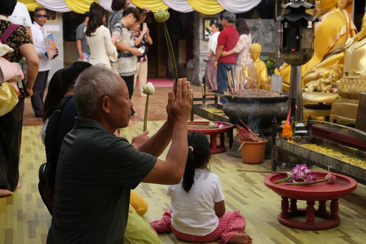 An older man kneels in a Buddhist temple, holding a white lotus blossom between his hands folded in prayer. He wears a dark polo shirt and a black backpack. Beside him, a small girl with long black hair, wearing a white T-shirt and a pink checkered skirt, sits on the floor. In front of them stand golden Buddha statues, a large incense vessel with burning incense sticks, and red offering bowls with orchids and other gifts. In the background, numerous visitors move through the temple area, decorated with yellow-and-white fabric banners. The atmosphere is marked by devotion and religious dedication, typical of a Thai Buddhist temple.
