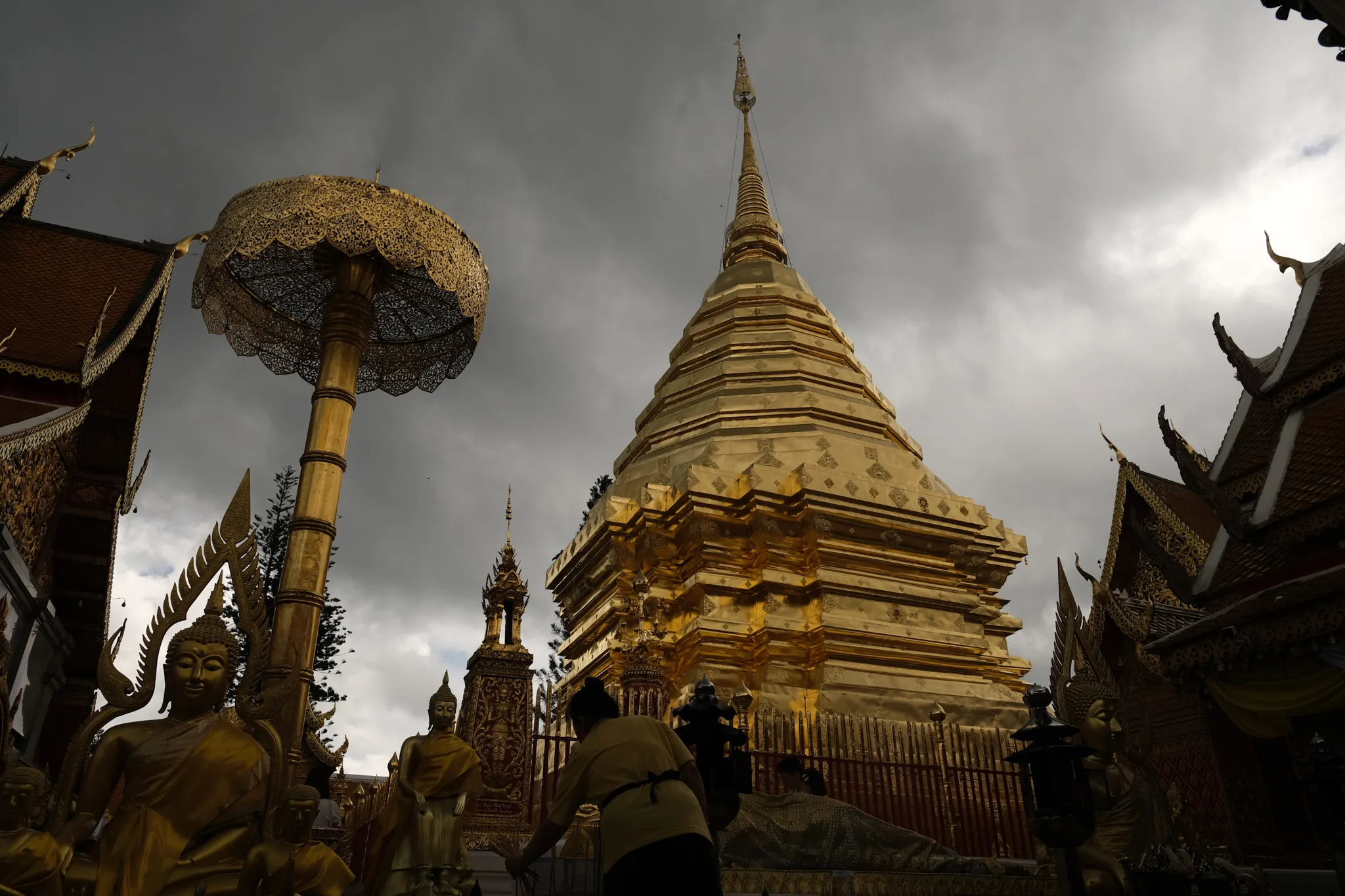 A towering golden stupa rises sharply toward a dark, cloud-filled sky, surrounded by richly decorated temple structures with intricate carvings and pointed rooflines. Several golden statues of serene seated and standing figures are arranged throughout the courtyard. A large ornate parasol stands prominently to the left, its delicate latticework catching the dim light. A few people move quietly near the base of the stupa, adding a sense of scale to the sacred space. The overall scene combines dramatic lighting with the elaborate details of traditional temple architecture.