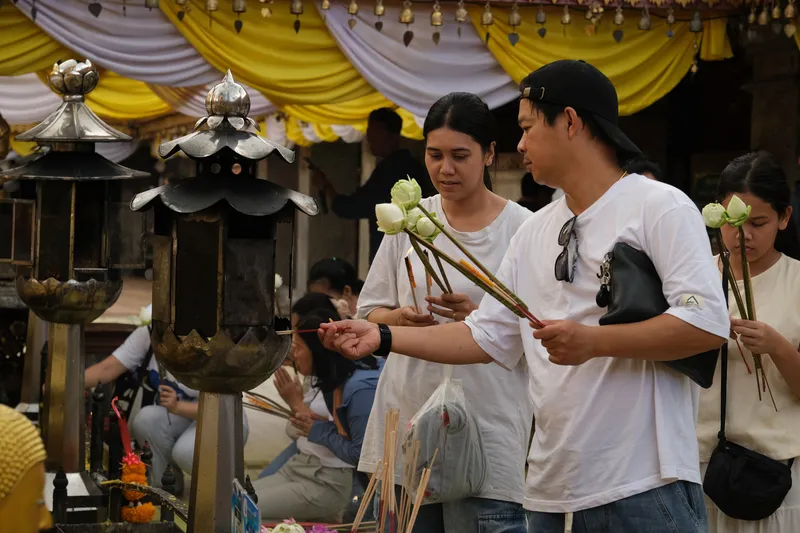 People stand at a shrine holding white lotus flowers and incense sticks, participating in a ritual offering. A man in a white shirt and cap reaches forward to light incense at a metal lantern-style structure, while others beside him prepare their offerings. More individuals are seen in the background praying with folded hands. Yellow and white drapery hangs above the area, and several ornate lanterns and flower arrangements surround the space, creating a solemn and respectful atmosphere.