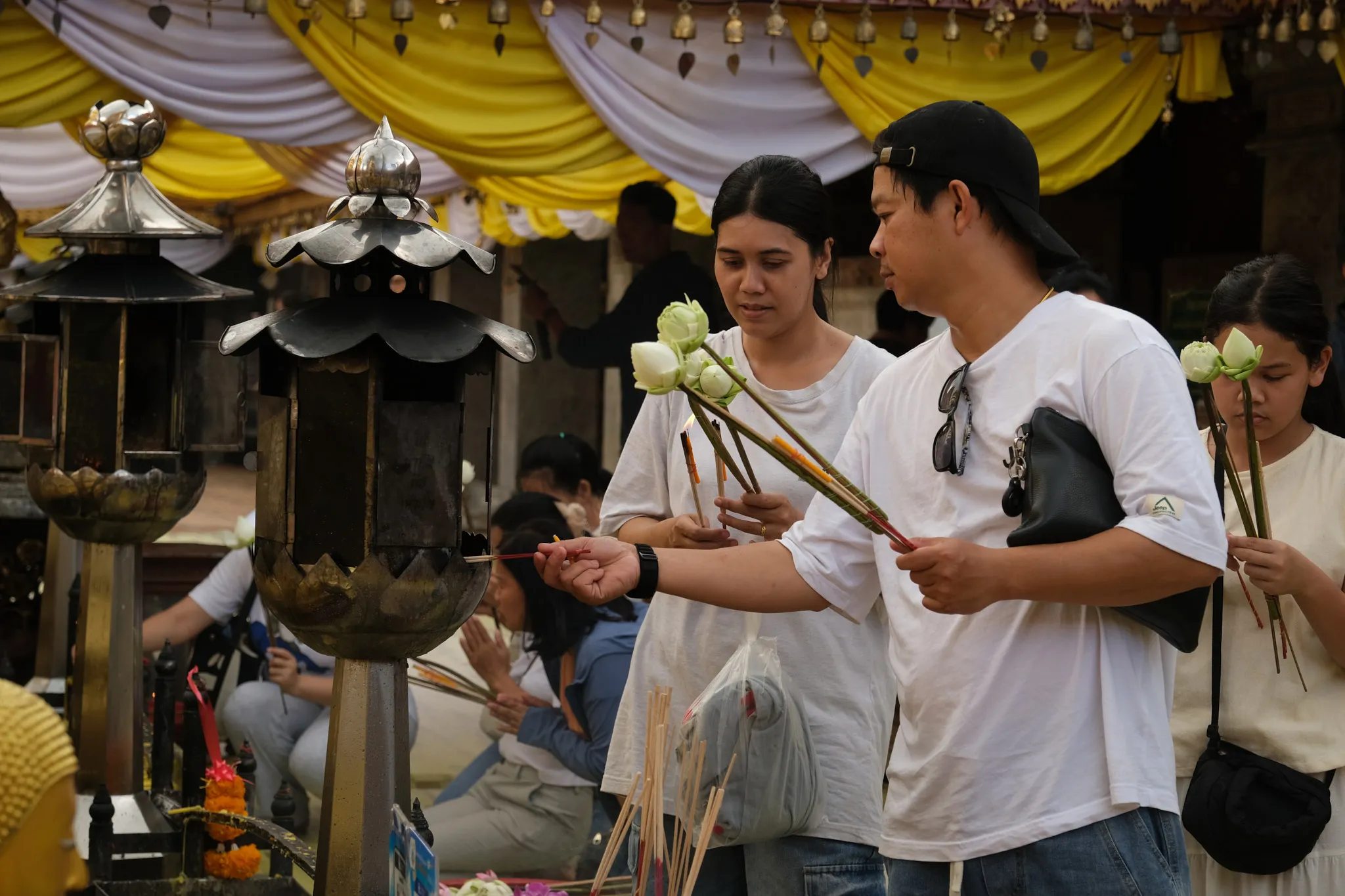 People stand at a shrine holding white lotus flowers and incense sticks, participating in a ritual offering. A man in a white shirt and cap reaches forward to light incense at a metal lantern-style structure, while others beside him prepare their offerings. More individuals are seen in the background praying with folded hands. Yellow and white drapery hangs above the area, and several ornate lanterns and flower arrangements surround the space, creating a solemn and respectful atmosphere.
