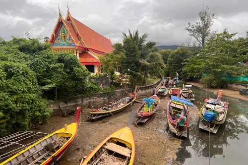 Several colorful longtail boats rest at low tide on the muddy shore of a narrow canal in Kamala, Kathu District, Phuket Province, Thailand. The traditional wooden Thai boats in yellow, red, blue, and green are moored to the bank with ropes and partly covered by tarps. In the background on the left rises a magnificent Buddhist temple (Wat) with a steep orange-red tiled roof and an elaborately decorated gable featuring golden carvings and a turquoise Buddha figure. Lush tropical vegetation with palms and densely foliaged trees surrounds the temple and lines both sides of the canal. The sky is covered by thick gray clouds, and the forested hills of Phuket can be seen in the background. The water in the canal is low, exposing large stretches of the muddy bottom.