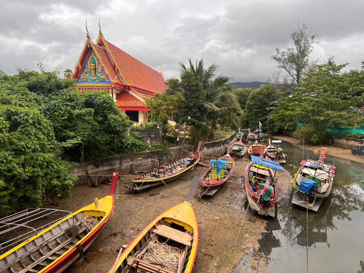 Several colorful longtail boats rest at low tide on the muddy shore of a narrow canal in Kamala, Kathu District, Phuket Province, Thailand. The traditional wooden Thai boats in yellow, red, blue, and green are moored to the bank with ropes and partly covered by tarps. In the background on the left rises a magnificent Buddhist temple (Wat) with a steep orange-red tiled roof and an elaborately decorated gable featuring golden carvings and a turquoise Buddha figure. Lush tropical vegetation with palms and densely foliaged trees surrounds the temple and lines both sides of the canal. The sky is covered by thick gray clouds, and the forested hills of Phuket can be seen in the background. The water in the canal is low, exposing large stretches of the muddy bottom.