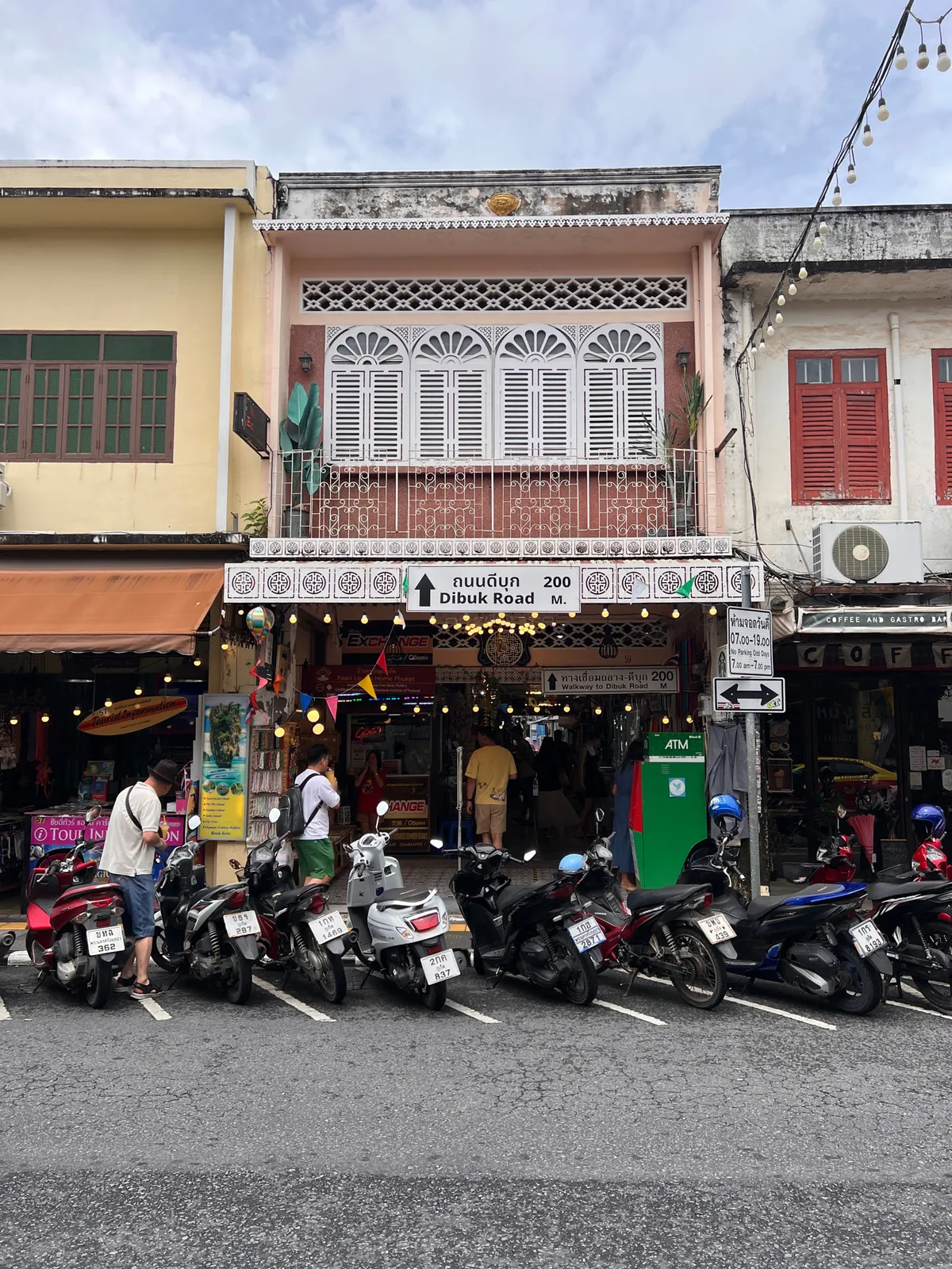 Street scene in the old town of Phuket (เมืองเก่าภูเก็ต), Thailand, with a row of Sino-Portuguese shophouse facades. At the center stands a pink building with ornate white shutters and fan-shaped decorations on the upper floor, along with a wrought-iron balcony featuring red decorative panels. Next to it on the left is a yellow building with green shutters and an orange awning, housing a travel agency labeled "Tour Information." To the right is a building with red shutters and a sign reading "Coffee and Gastro Bar." In front of the buildings, numerous scooters are parked in a row. A green street sign points to Dibuk Road 200 meters away, next to a notice about the "Walkway to Dibuk Road." A green ATM stands at the curb. Several pedestrians are visible, including a man in a hat and white T-shirt and another with a backpack. Strings of lights stretch between the buildings, and the sky is partly cloudy. The scene conveys the lively, tourist-oriented atmosphere of Phuket's historic old town.