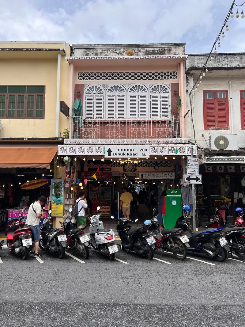 A row of parked scooters lines a wet street in front of a lively storefront beneath a pastel-colored shophouse with ornate white shutters and decorative balcony railings. Several people browse outside the shop, which displays bright signage, hanging lights, and an ATM on the right side. Adjacent buildings feature colorful facades with green and red window frames, creating a vibrant streetscape under a cloudy sky.