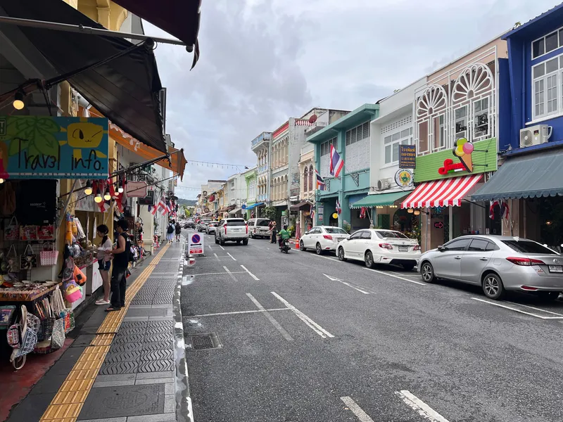 Busy street lined with colorful shophouses featuring decorative facades, awnings, and small storefronts. Several parked cars stretch along the right side while a few people browse goods at a shop on the left. Flags hang from many of the buildings, and the wet pavement reflects the cloudy sky above. Overhead string lights cross the street in the distance, adding to the lively atmosphere of this historic urban district.