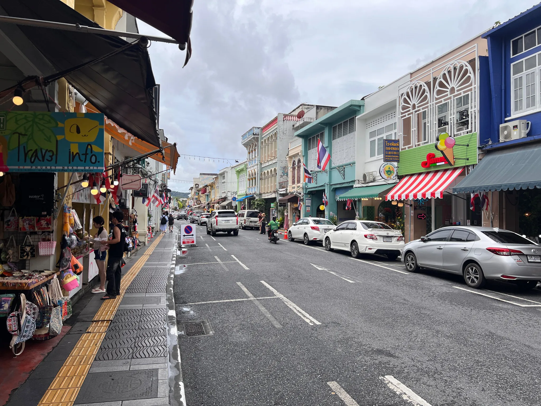 Busy street lined with colorful shophouses featuring decorative facades, awnings, and small storefronts. Several parked cars stretch along the right side while a few people browse goods at a shop on the left. Flags hang from many of the buildings, and the wet pavement reflects the cloudy sky above. Overhead string lights cross the street in the distance, adding to the lively atmosphere of this historic urban district.