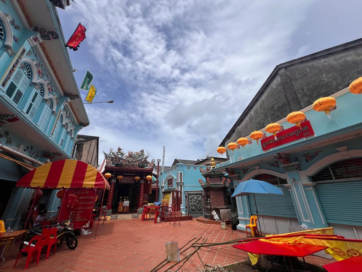 Courtyard of a Chinese temple in the old town of Phuket (เมืองเก่าภูเก็ต), Thailand, surrounded by pastel-blue Sino-Portuguese buildings with elaborate ornamentation and stucco work. At the center rises a traditional Chinese shrine with a colorful roof decorated with dragons and figures. Orange lanterns hang in a row between the buildings, along with a red sign in Thai script. On the red-tiled floor stand red plastic chairs, a motorbike, a large bronze incense vessel, and red-and-yellow striped parasols and awnings. Colorful flags in red, green, and yellow flutter on the facade of the left building. A blue parasol stands next to the shrine. The sky is partly cloudy with bright cloud formations breaking through.